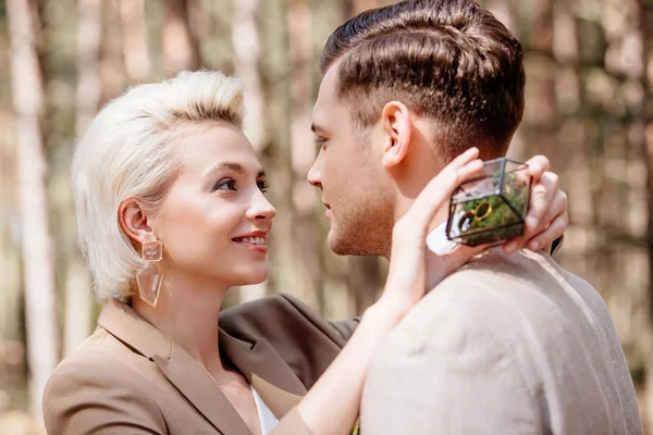 Lächelndes blondes Mädchen hält Box mit Eheringen und umarmt Bräutigam im Wald — Stockfoto