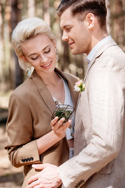 Sonriente novio y novia caja de espera con anillos de boda en el bosque - foto de stock