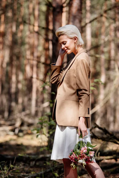 Femme blonde souriante en veste tenant bouquet en forêt — Photo de stock
