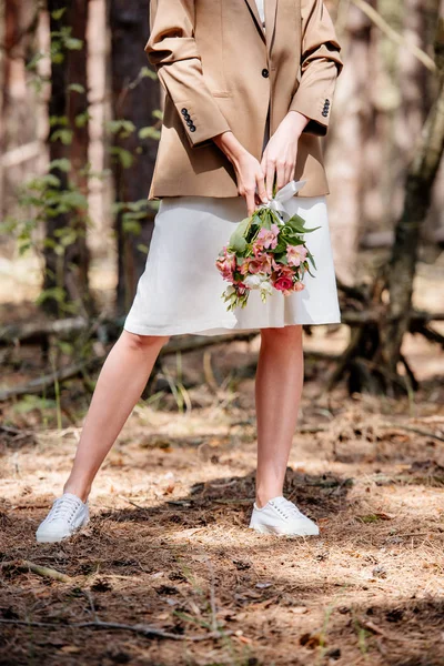 Cropped view of bride in brown jacket holding wedding bouquet in forest — Stock Photo
