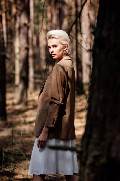 Elegant serious blonde woman looking at camera while standing in forest — Stock Photo