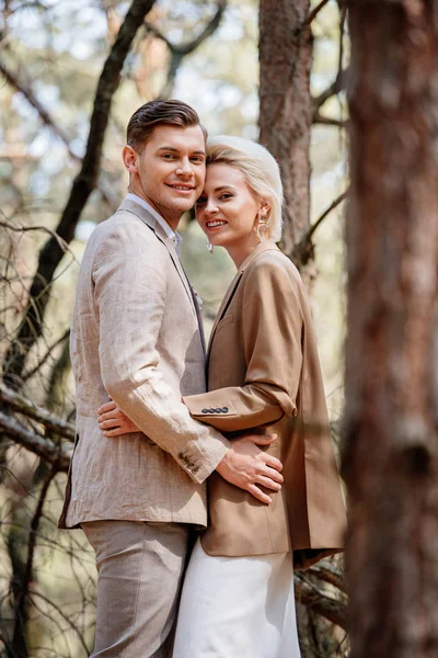Стильная улыбающаяся пара в элегантных куртках, обнимающихся в лесу — стоковое фото
