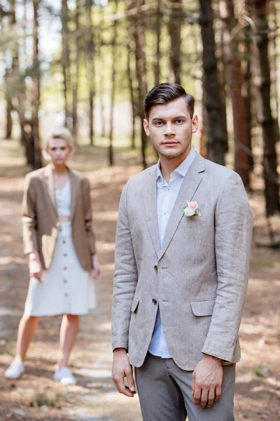 Stylish groom with boutonniere on jacket and bridegroom standing in forest and looking at camera — Stock Photo