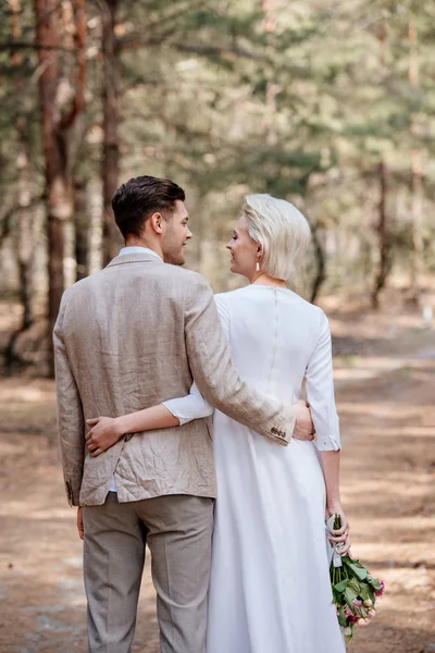 Vista trasera de la pareja recién casada abrazándose y mirándose en el bosque - foto de stock