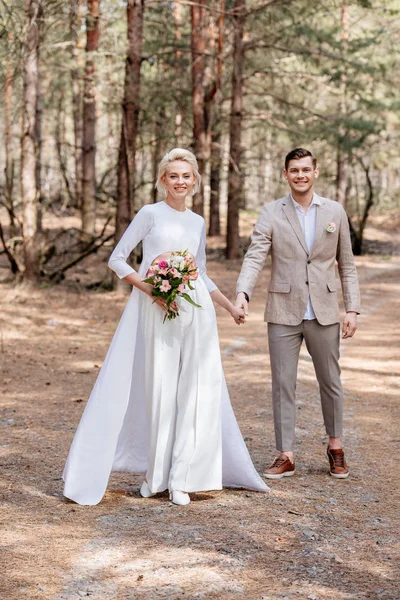 Full length view of smiling just married couple holding hands in forest — Stock Photo