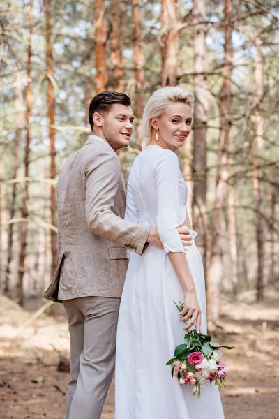 Smiling bridegroom in formal wear embracing bride in forest — Stock Photo