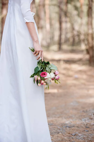 Cropped view of bride in white attire holding wedding bouquet in forest — Stock Photo