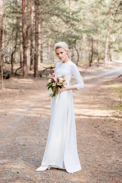 Visão de comprimento total da bela noiva segurando buquê de casamento na floresta — Fotografia de Stock