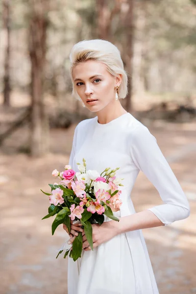 Atractiva novia rubia celebración de ramo de bodas en el bosque - foto de stock