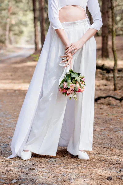 Vista cortada da noiva em traje branco segurando buquê de casamento na floresta — Fotografia de Stock