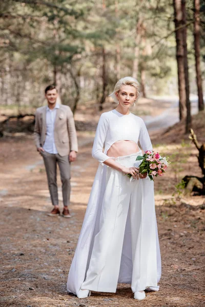 Vue pleine longueur de la mariée avec bouquet de fleurs et marié dans la forêt — Photo de stock