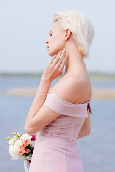 Задумчивая блондинка в розовом платье держит букет — стоковое фото