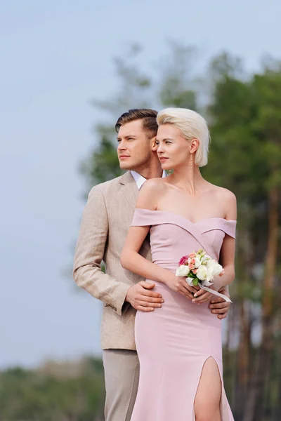 Casal elegante com buquê de casamento abraçando e olhando para longe — Fotografia de Stock