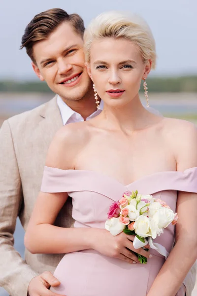 Happy just married couple with wedding bouquet looking at camera — Stock Photo