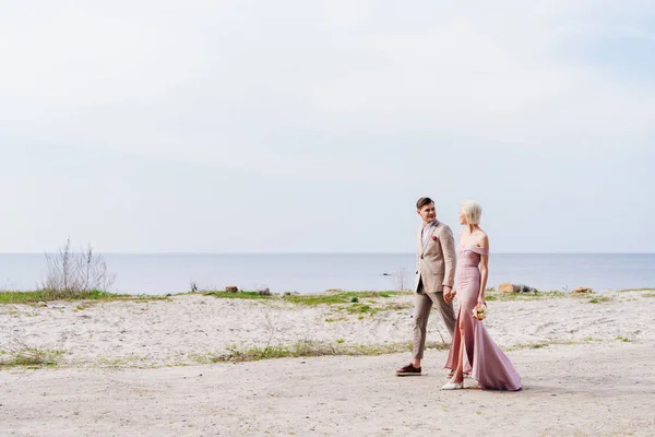 Vista completa de pareja recién casada cogida de la mano y caminando por la costa - foto de stock