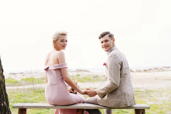 Sonriente pareja recién casada sentada en el banco y cogida de la mano - foto de stock
