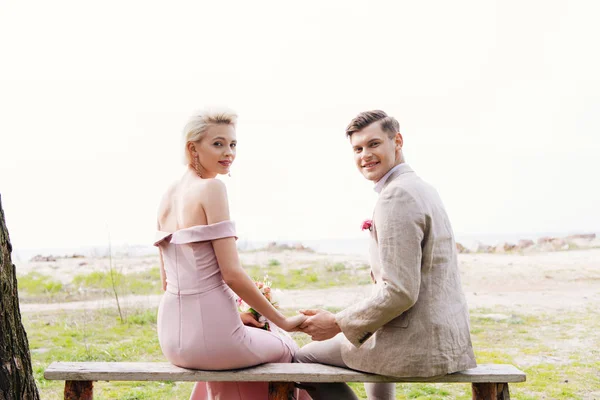Smiling just married couple sitting on bench and holding hands — Stock Photo