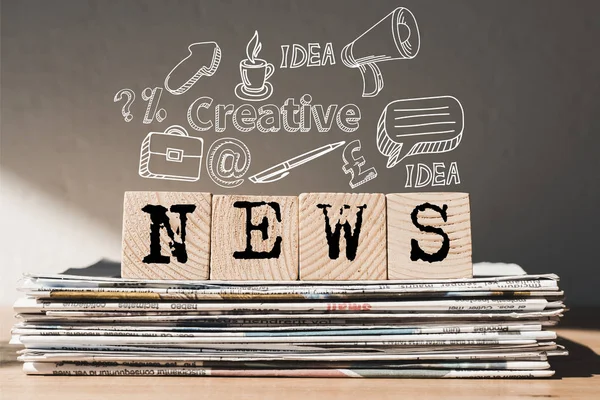 Holzblöcke mit schwarzem Nachrichtenschriftzug auf Zeitungsstapel mit Ideensymbolen oben — Stockfoto