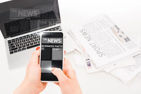 Selective focus of woman holding smartphone with business news illustration near laptop and newspapers — Stock Photo