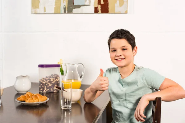 Niño sonriente sentado en la mesa durante el desayuno y mostrando el pulgar hacia arriba - foto de stock