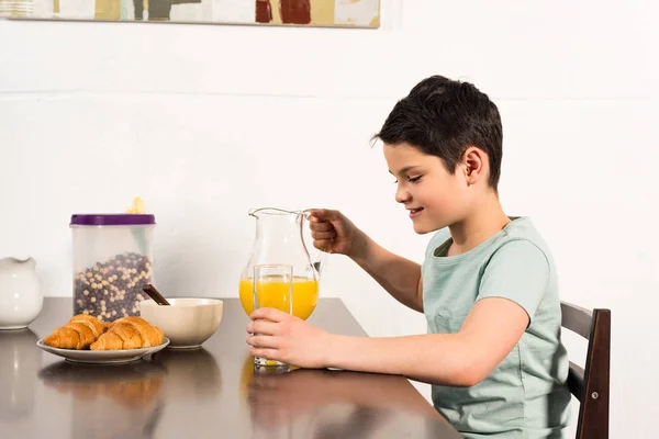 Ragazzo sorridente che tiene in mano vetro e brocca con succo d'arancia in cucina — Foto stock