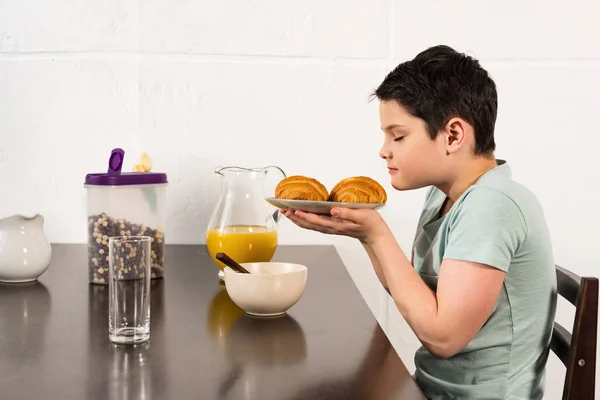 Niño oliendo croissants con los ojos cerrados durante el desayuno en la cocina - foto de stock