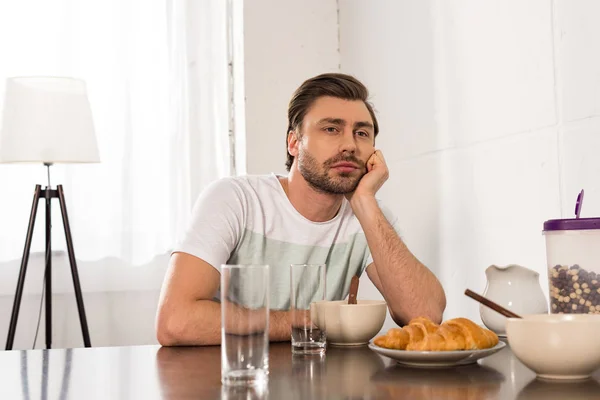 Nachdenklicher Mann sitzt am Tisch und stützt beim Frühstück in der Küche das Gesicht mit der Hand ab — Stockfoto