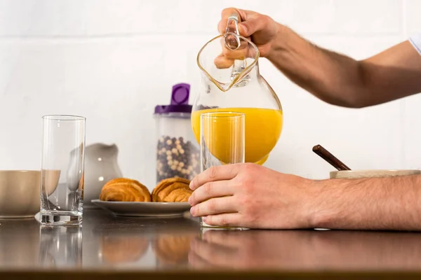 Cropped view of man pouring orange juice in glass during breakfast in kitchen — Stock Photo