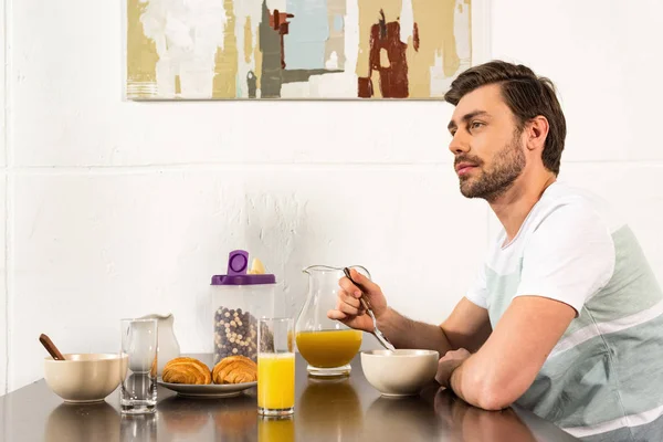 Hombre barbudo sentado en la mesa durante el desayuno y soñando mirando hacia otro lado en la cocina - foto de stock