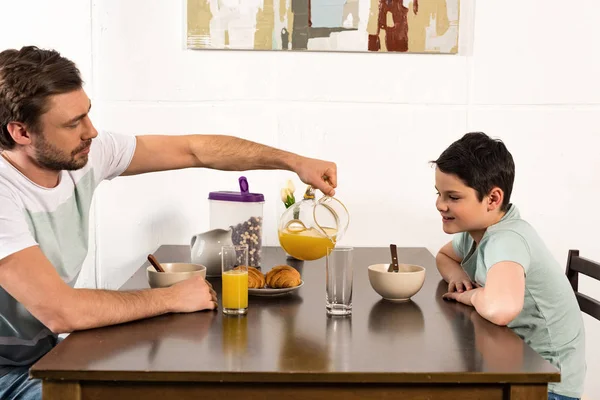 Barbudo papá verter jugo de naranja a hijo durante el desayuno - foto de stock