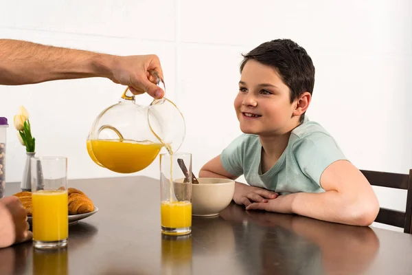 Vista recortada de papá vertiendo jugo de naranja al hijo durante el desayuno - foto de stock