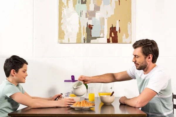 Pai derramando leite na tigela durante o café da manhã com o filho — Fotografia de Stock