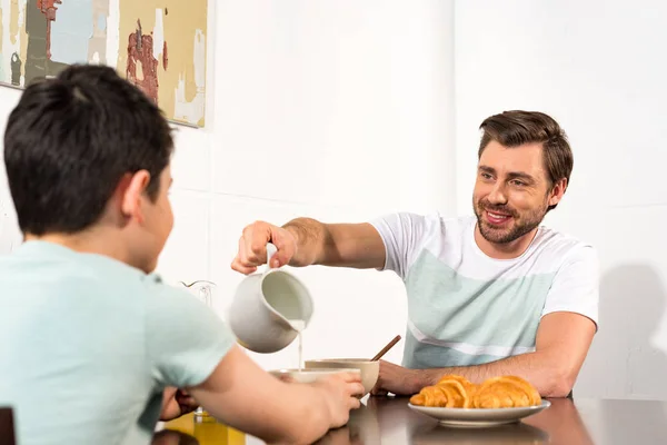 Papá vertiendo leche en un tazón durante el desayuno con su hijo - foto de stock