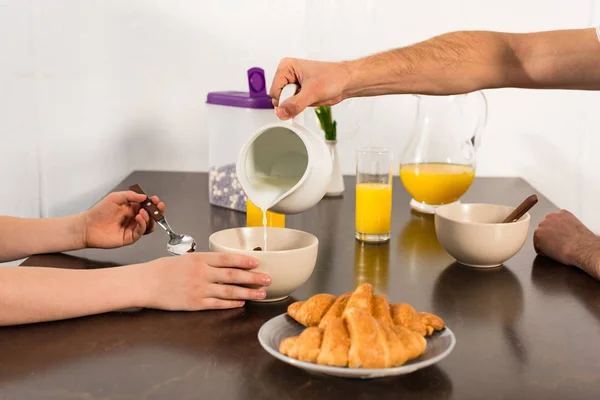 Vista recortada de papá vertiendo leche en un tazón durante el desayuno con su hijo - foto de stock