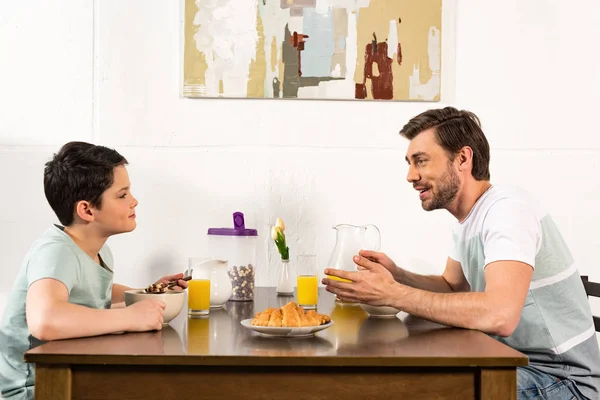 Sorrindo pai e filho tomando café da manhã e olhando um para o outro na cozinha — Fotografia de Stock