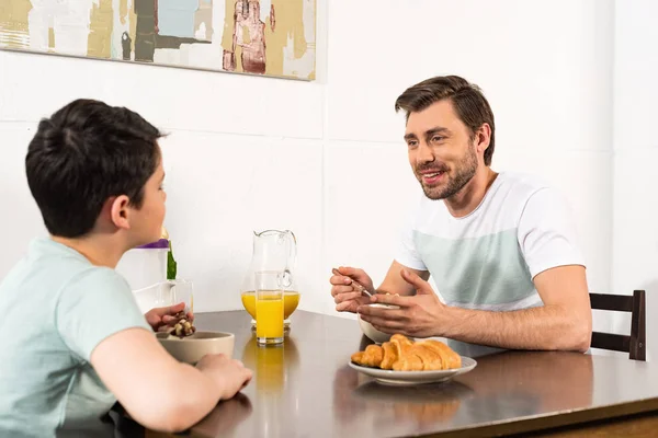 Sorrindo pai e filho tomando café da manhã e olhando um para o outro na cozinha — Fotografia de Stock