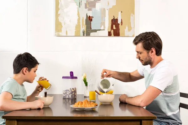 Pai derramando leite na tigela enquanto o pecado beber suco de laranja durante o café da manhã — Fotografia de Stock