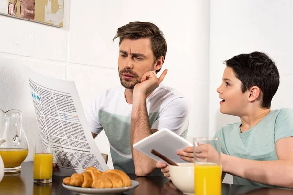 Pai focado ler jornal e filho usando tablet digital durante o café da manhã — Fotografia de Stock