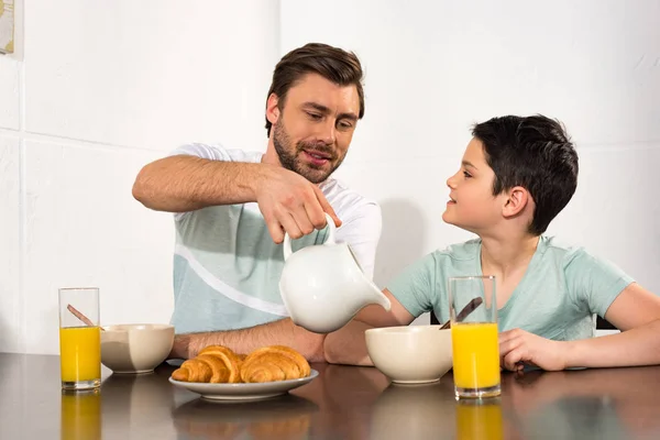 Sonriente papá verter leche a hijo durante el desayuno en la cocina - foto de stock