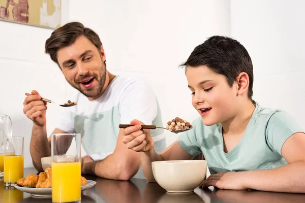 Sonriente padre e hijo comiendo cereal de desayuno en la cocina - foto de stock