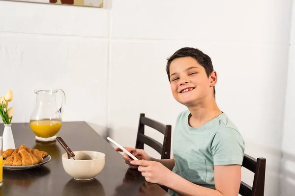 Menino sorrindo usando tablet digital durante o café da manhã na cozinha — Fotografia de Stock