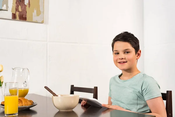 Garçon souriant utilisant tablette numérique pendant le petit déjeuner dans la cuisine — Photo de stock