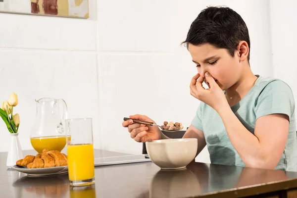 Niño pellizcando la nariz mientras come cereales de desayuno en la cocina - foto de stock