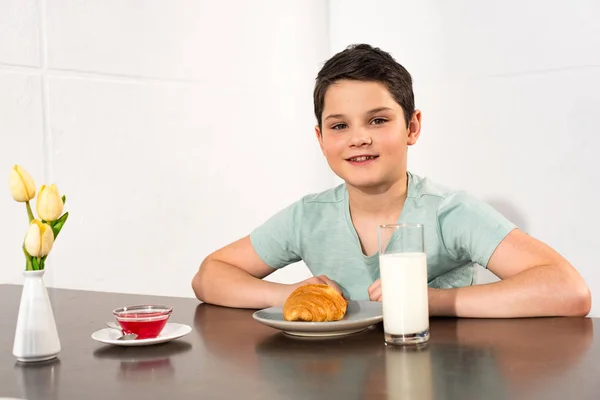 Niño sonriente sentado en la mesa con croissant, jarabe y vaso de leche - foto de stock