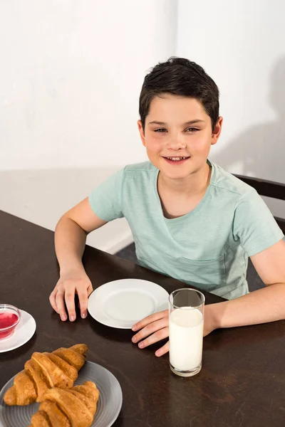 Вид сверху на улыбающегося мальчика, сидящего за столом с круассанами, сиропом и стаканом молока — стоковое фото