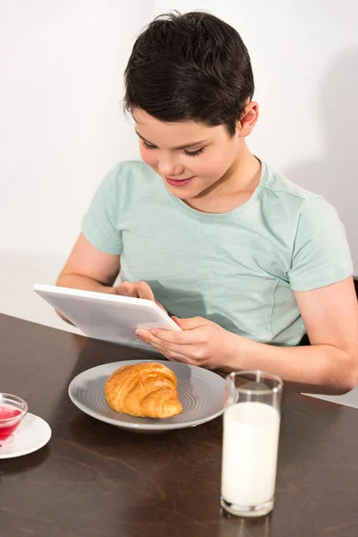 Улыбающийся мальчик с цифровым планшетом во время завтрака на кухне — стоковое фото