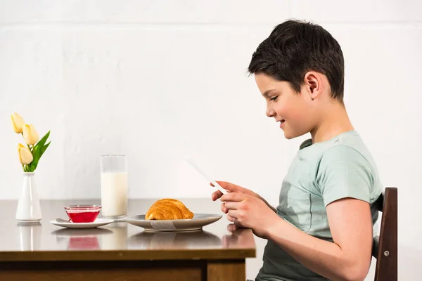 Vue latérale du garçon souriant en utilisant une tablette numérique pendant le petit déjeuner dans la cuisine — Photo de stock