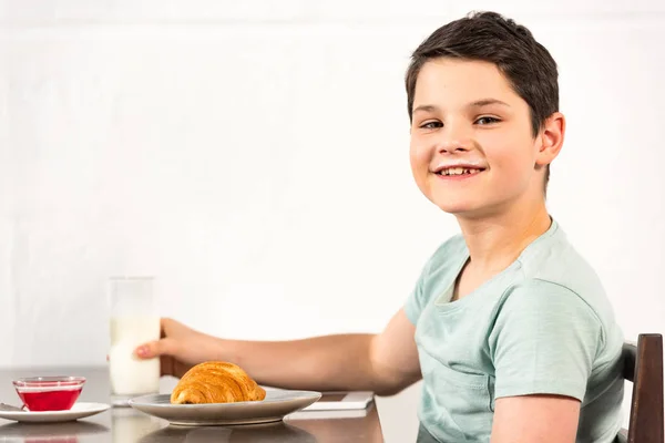 Niño sonriente sentado en la mesa con croissant, jarabe y vaso de leche - foto de stock