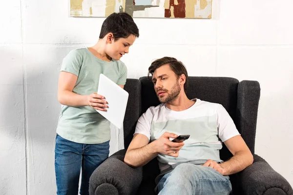 Son showing digital tablet to father sitting in armchair with remote controller — Stock Photo
