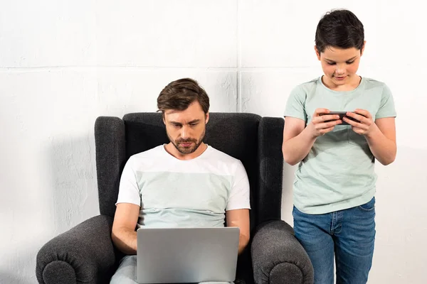 Сын пользуется смартфоном, а отец ноутбуком в гостиной — стоковое фото